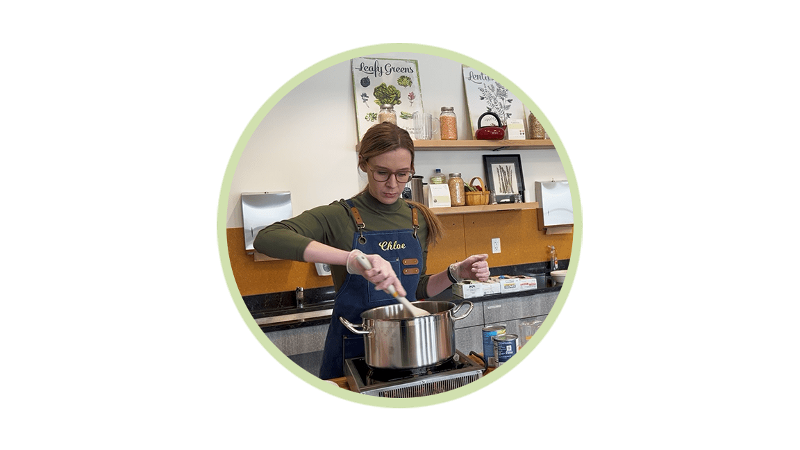Woman wearing a blue apron labeled Chloe stirs a pot on a stove in a kitchen.