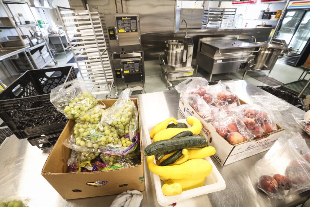 Commercial kitchen prep area with stainless steel counters and racks; boxes of grapes, bananas, and squash in foreground.