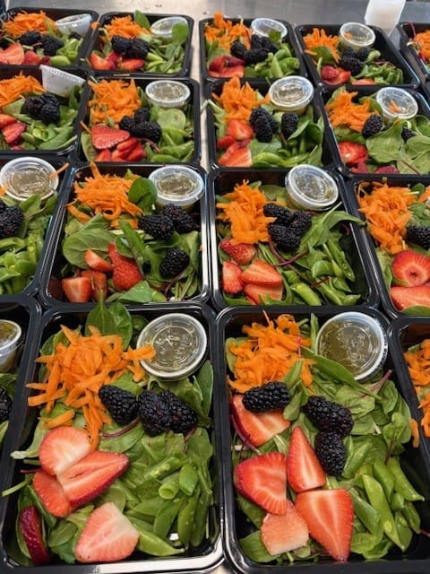 Multiple meal-prep salad containers filled with leafy greens, sliced strawberries, blackberries, shredded carrots, and a side dressing.