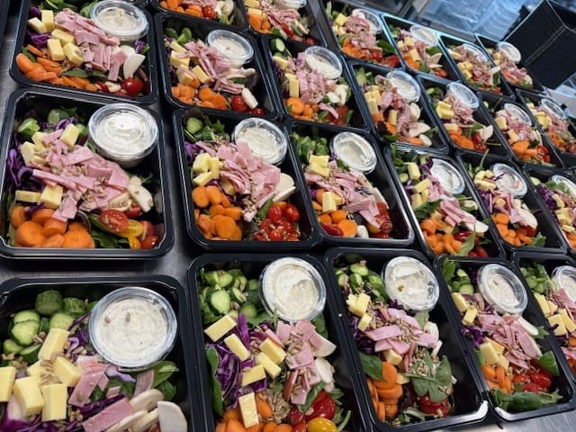 Rows of black lunch salad boxes with mixed greens, ham, cheese, carrots, cucumbers, tomatoes, and a cup of dressing as garnish.