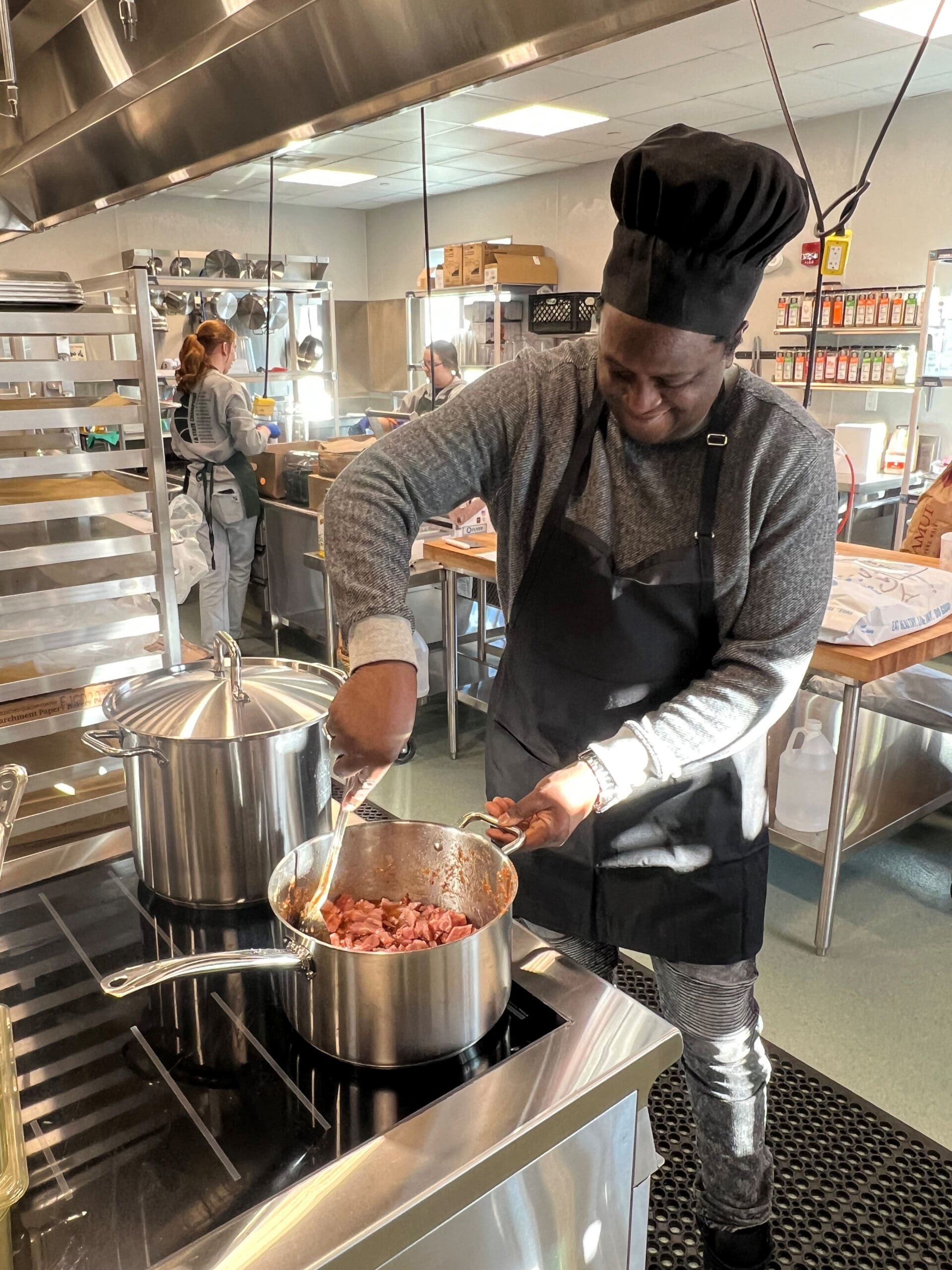Chef in a black toque and apron stirs diced meat in a large stainless pot on a commercial stove, with teammates working in the background.