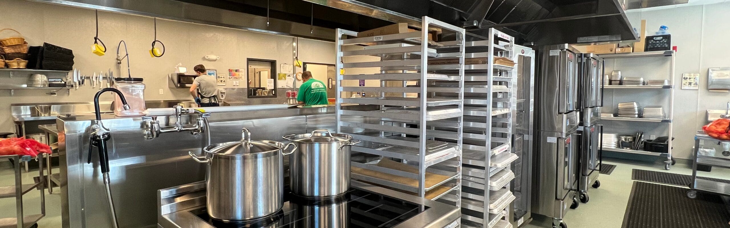Commercial kitchen with stainless steel appliances, two large pots on a stovetop, and tall racks of baking sheets in the background.