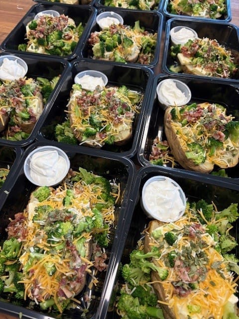 Rows of takeout taco bowls in black trays, topped with cilantro, shredded cheese, greens, and a side cup of sour cream.