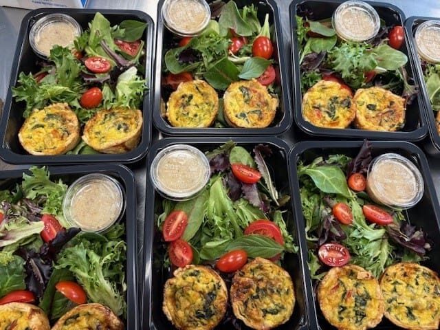 Top-down view of six meal-prep boxes with greens, cherry tomatoes, and mini quiches, plus a cup of dressing.