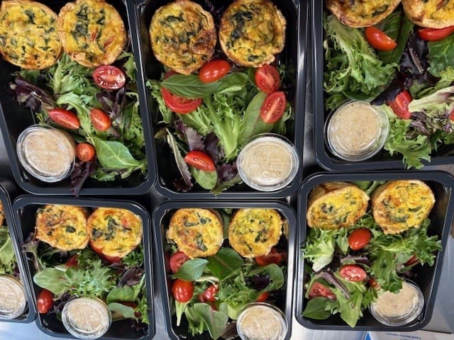 Top-down view of six meal-prep boxes with greens, cherry tomatoes, and mini quiches, plus a cup of dressing.