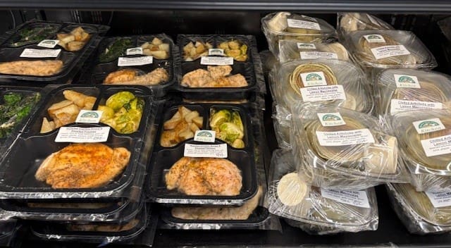 Assorted ready-to-eat meals in black plastic trays with chicken and vegetables on a supermarket shelf.