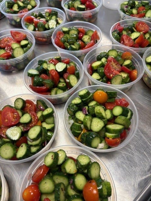 Clear bowls filled with cucumber and tomato salad (red onions visible) arranged on a stainless steel table.