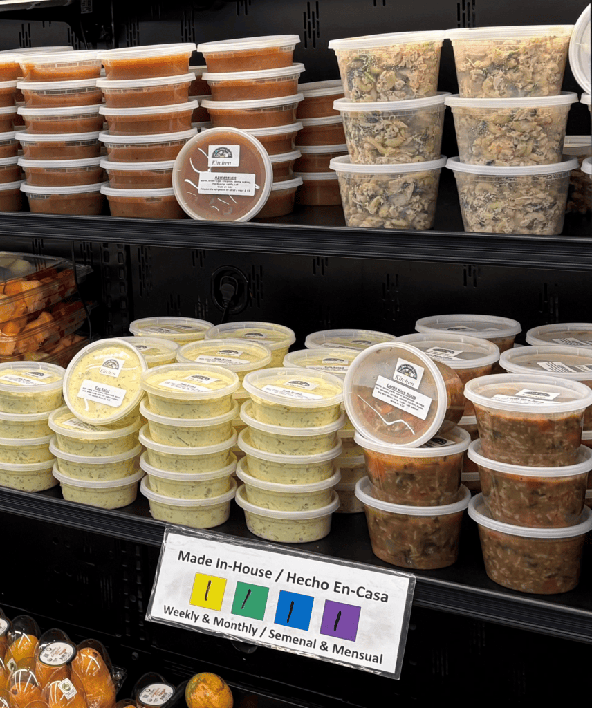 Shelf display of homemade meals and sauces in clear tubs, orange sauce on left and mixed pasta dishes on right, with a 'Made In-House / Hecho En-Casa' sign visible below.