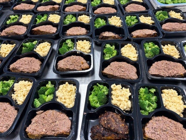 Overhead view of many black meal-prep containers in a grid, each with ground beef, broccoli, and scrambled eggs.