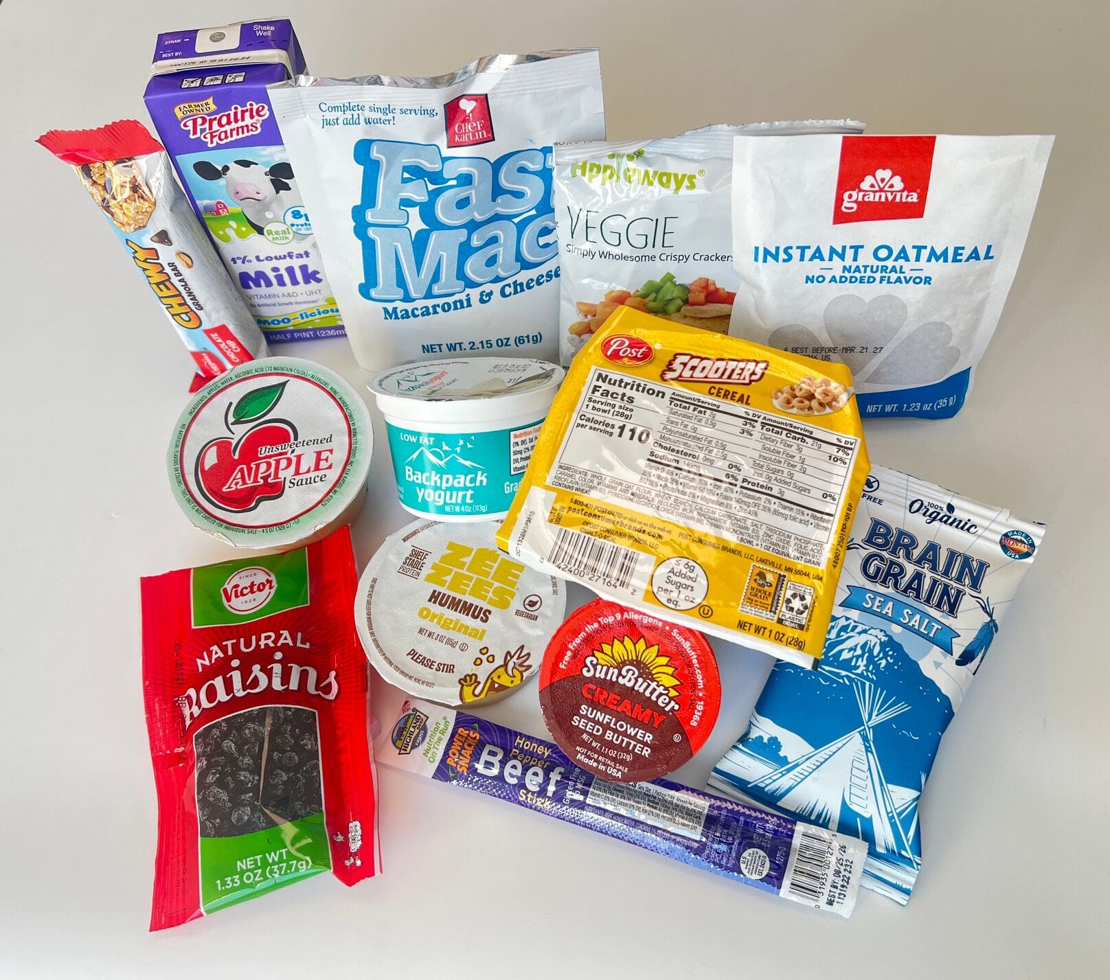Assortment of packaged snacks and breakfast items, including oats, granola, crackers, and yogurt cups arranged on a white surface.