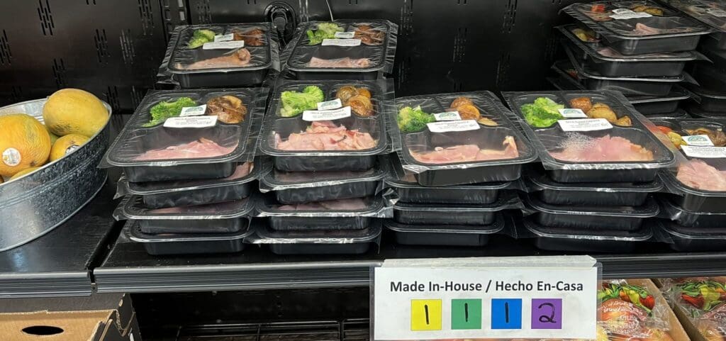 Rows of black plastic meal trays with ham and broccoli on a store shelf, labels visible on each tray.